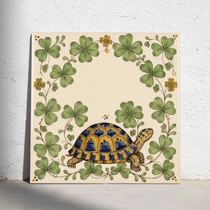 May include: A square ceramic tile featuring a central illustration of a tortoise with a yellow and blue patterned shell. The tortoise is surrounded by a decorative border of green clover leaves and small yellow flowers on a cream background.