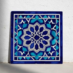 Könnte beinhalten: Quadratische Keramikfliese mit einem geometrischen Sternmuster in Blau- und Weißtönen. Die Fliese hat einen dunkelblauen Rand und ein sich wiederholendes Muster aus Dreiecken und Rauten. Das Design ist auf einem weißen Hintergrund zentriert.