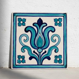 May include: Square ceramic tile with a floral design in shades of teal and blue. The central motif is a stylized flower, surrounded by four smaller floral accents and a border. The tile is set against a white background.