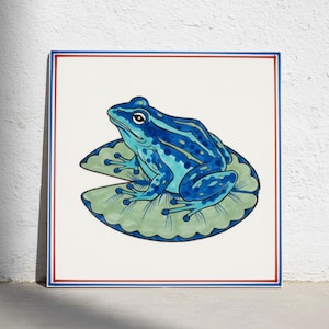 Puede incluir: Un azulejo de cerámica cuadrado con una ilustración pintada a mano de una rana azul brillante con manchas azul oscuro sentada sobre un nenúfar verde claro. El azulejo tiene un fondo blanco y un borde fino de líneas rojas y azules.