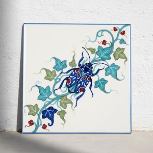 May include: A square ceramic tile featuring a central blue beetle with white and red patterned markings. The beetle is surrounded by a winding vine with light green and blue leaves and small red berries, all set against a plain white background.