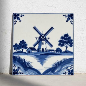 Può includere: Piastrella quadrata in ceramica con un design a mulino a vento blu e bianco. L'immagine raffigura un mulino a vento, alberi e acqua, con decorazioni floreali agli angoli. Lo stile ricorda la ceramica di Delft, uno stile ceramico classico.