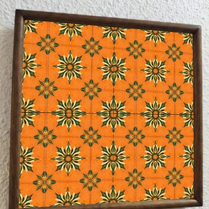 May include: A framed artwork with a repeating floral tile pattern. The tiles are orange with green and cream floral designs. The frame is dark brown wood, contrasting with the vibrant tile design. The artwork is a decorative piece.