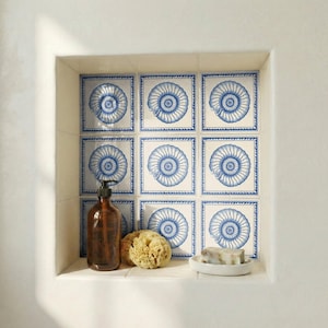 May include: A bathroom niche features nine square tiles with a blue nautilus shell design. A brown soap dispenser, a natural sponge, and a bar of soap on a white dish are on the shelf below the tiles.