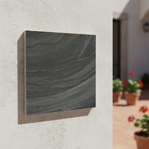 May include: A square, dark gray outdoor wall tile with a textured, layered pattern. The tile is mounted on a white stucco wall, casting a shadow. The tile's surface has a natural, stone-like appearance, suitable for exterior design.