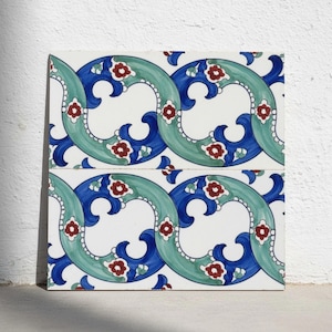 May include: A square ceramic tile featuring a repeating pattern of interlocking teal and dark blue curved bands on a white background. The design is accented with small red and white floral motifs arranged along the flowing, wave-like shapes.