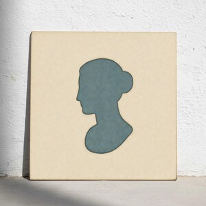 Puede incluir: Una obra de arte cuadrada beige con una silueta azul del perfil de una mujer con un moño. La obra tiene una apariencia texturizada y está sobre un fondo blanco.