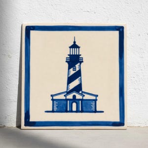 Puede incluir: Azulejo cerámico cuadrado con una ilustración de faro azul y blanco. El faro, con su torre a rayas, está centrado sobre un fondo crema, enmarcado por un borde azul. Adecuado para la decoración del hogar.