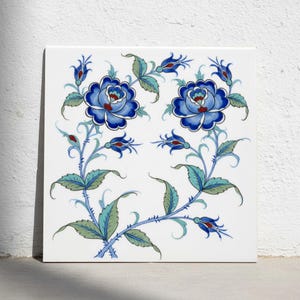 Könnte beinhalten: Eine quadratische weiße Keramikfliese mit einem handgemalten Blumenmuster. Das Design zeigt zwei große, symmetrische blaue Rosen mit detaillierten Blütenblättern, umgeben von hellgrünen Blättern, dornigen Stielen und kleinen blauen Blütenknospen.