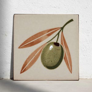 Puede incluir: Azulejo cerámico cuadrado con un diseño de aceituna verde y rama de olivo. La aceituna es verde con un hueso marrón oscuro, y las hojas son de color marrón rojizo. El azulejo tiene un fondo color crema.