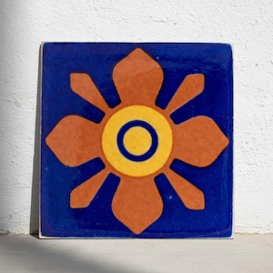May include: Square ceramic tile with a floral design. The tile has a dark blue background with an orange, yellow, and blue flower. The flower has a central yellow circle with a blue ring.