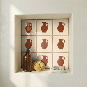 Puede incluir: Estantería empotrada con azulejos de jarras de color terracota. Un dispensador de jabón de vidrio marrón, una esponja natural y un plato blanco pequeño con jabón están en el estante. Los azulejos tienen un patrón de jarra repetido.