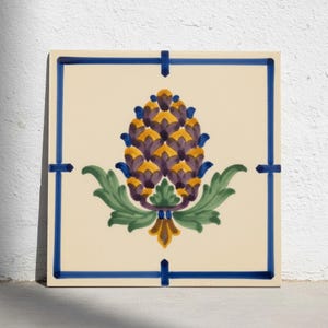 Puede incluir: Un azulejo de cerámica cuadrado con fondo color crema que presenta un motivo central pintado a mano de una piña estilizada en tonos morado, amarillo y verde. El diseño está enmarcado por un borde azul fino con pequeños acentos.