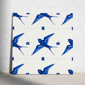 Op de afbeelding: Een vierkant raster van negen keramische tegels met een herhalend patroon van blauwe zwaluwen geschilderd op een witte achtergrond. Elke tegel toont een gestileerde vogel in vlucht met gespreide vleugels, wat een decoratief geheel vormt.