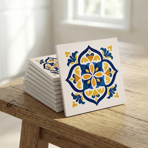 Puede incluir: Una pila de posavasos cuadrados de cerámica con un diseño floral. Los posavasos tienen un fondo blanco con un patrón simétrico en tonos azules y amarillos. Se muestran sobre una superficie de madera.