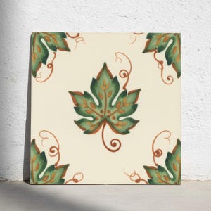 May include: A square ceramic tile with an off-white background featuring a central green grape leaf design with orange veins and tendrils. The four corners of the tile are decorated with matching green leaf patterns and orange swirling vine accents.