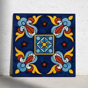 May include: Square ceramic tile with a vibrant, symmetrical design. The tile features a deep blue background with a central square and four repeating floral-like motifs in yellow, red, and light blue. Small black flower accents complete the design.