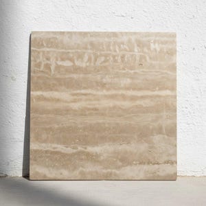Puede incluir: Una baldosa cuadrada de piedra beige con un patrón natural en capas. La baldosa tiene un color marrón claro con vetas más oscuras y sutiles variaciones en la textura, lo que sugiere un material de piedra natural. La baldosa está apoyada contra una pared blanca.