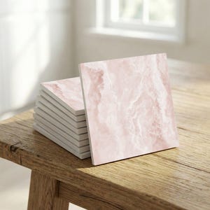 Op de afbeelding: Een stapel vierkante decoratieve tegels met een roze marmerontwerp. De tegels zijn gestapeld op een houten oppervlak, met één tegel die tegen de stapel leunt. De tegels hebben een witte rand en een lichtroze, wervelend patroon.