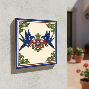 May include: Square decorative tile with a cream background, featuring two blue birds with red chests, facing each other above a red and green floral design. The tile has a blue border and green corner accents. Mounted on a white wall.