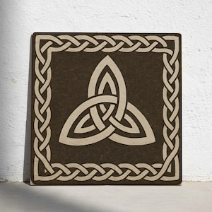 Może przedstawiać: Kwadratowa, brązowa płytka dekoracyjna z beżowym wzorem węzła celtyckiego. Węzeł to symbol triquetry, otoczony beżową, plecioną obwódką. Płytka opiera się o białą ścianę.