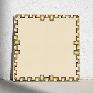 Può includere: Una piastrella quadrata in ceramica con un centro color crema e un bordo decorativo. Il bordo presenta un motivo geometrico greco ripetitivo dipinto in tonalità alternate di blu, giallo e verde, creando una cornice strutturata.