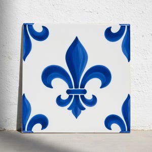 May include: A square ceramic tile featuring a central blue fleur-de-lis design on a white background. The corners of the tile are decorated with matching blue curved leaf-like motifs, creating a symmetrical pattern across the flat surface.