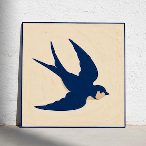 May include: Wooden wall art with a navy blue swallow design on a cream background. The bird is depicted in flight, with its wings spread. The artwork has a minimalist style and is framed with a dark blue border. The dimensions are approximately 20 cm x 20 cm.