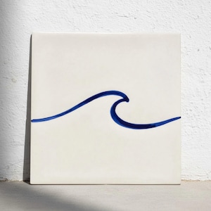 Puede incluir: Azulejo cerámico blanco cuadrado con un diseño de ola azul pintado a mano. La ola es una ilustración sencilla y estilizada, que añade un toque de decoración costera. El azulejo es limpio y moderno, adecuado para diversas aplicaciones de decoración del hogar.