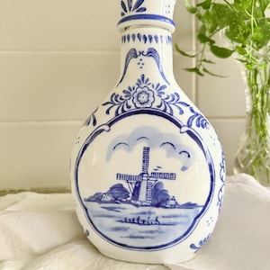 May include: A white ceramic bottle with a blue hand-painted design featuring a central windmill scene within an oval frame. The bottle has a narrow neck, a flared top, and decorative blue floral patterns surrounding the main illustration.