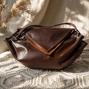 Puede incluir: Un bolso de mano de cuero marrón oscuro con una solapa frontal triangular y un borde de cuero marrón más claro. El bolso tiene una estructura suave y holgada con correas de cuero finas que pasan por ojales metálicos laterales.