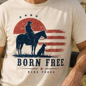 Op de afbeelding: Ivoor t-shirt met een cowboy te paard en een hond, tegen een rood-blauwe grafische achtergrond die op een vlag lijkt. De tekst "BORN FREE" en "RIDE PROUD" staan eronder gedrukt. Het shirt is gelabeld "IVORY".