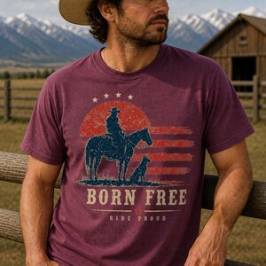 Op de afbeelding: Bessenkleurig T-shirt met een afbeelding van een cowboy op een paard, een hond en de tekst "BORN FREE". Het ontwerp bevat een rode en blauwe zonsondergang en sterren. Het shirt wordt gedragen met een cowboyhoed.