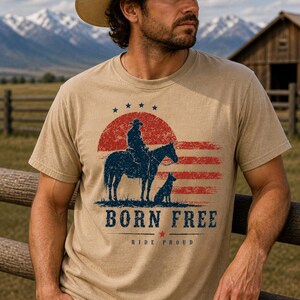 Op de afbeelding: T-shirt in zandkleur met een afbeelding van een cowboy te paard, een hond en de tekst "BORN FREE" en "RIDE PROUD". Het ontwerp bevat rode en blauwe elementen. Het woord "SANDSTONE" staat onderaan.