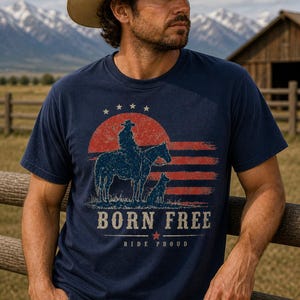 Op de afbeelding: Marineblauw T-shirt met een afbeelding van een cowboy op een paard, een hond en de tekst "BORN FREE RIDE PROUD". Het ontwerp bevat een rood-witte gestreepte achtergrond en een rode zon. Het woord "NAVY" staat onderaan.