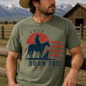 Op de afbeelding: Mosgroen t-shirt met een afbeelding van een cowboy te paard, een hond en de tekst "BORN FREE". Het ontwerp bevat een rode en blauwe zonsondergang en sterren. Het model draagt een cowboyhoed en een blauwe spijkerbroek.