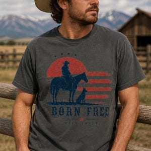 Op de afbeelding: Donkergrijs t-shirt met een cowboy-afbeelding. Het ontwerp toont een cowboy op een paard met een hond, een rood-blauwe zonsondergang en de tekst "BORN FREE". Het shirt wordt gedragen met een cowboyhoed en een spijkerbroek.