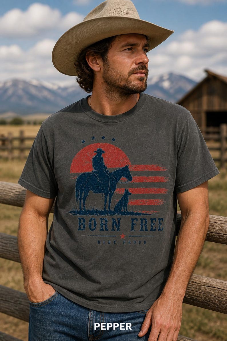 Op de afbeelding: Donkergrijs t-shirt met een cowboy-afbeelding. Het ontwerp toont een cowboy op een paard met een hond, een rood-blauwe zonsondergang en de tekst "BORN FREE". Het shirt wordt gedragen met een cowboyhoed en een spijkerbroek.