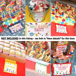 May include: A circus-themed party food table with various snacks, including popcorn, lollipops, pretzels, and fruit. The table is decorated with red and white striped paper and colorful polka dots. The signs on the table read "Lollipops", "Popcorn", "Pretzels", and "Salt Water Taffy".