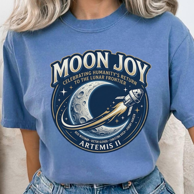May include: A light blue t-shirt featuring a circular graphic with the words "MOON JOY" and "CELEBRATING HUMANITY'S RETURN TO THE LUNAR FRONTIER." The graphic includes a crescent moon, a rocket, and the text "ARTEMIS II."