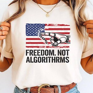 May include: Cream-colored t-shirt featuring a graphic design. The design includes a stylized American flag, a robotic hand pointing, and the text "FREEDOM. NOT ALGORITHMS" in bold, black letters. The shirt is a casual wear item.