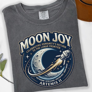 May include: A gray Comfort Colors t-shirt featuring a circular graphic with the words "MOON JOY" and "ARTEMIS II". The graphic depicts a crescent moon, a rocket, and the text "CELEBRATING HUMANITY'S RETURN TO THE LUNAR FRONTIER".