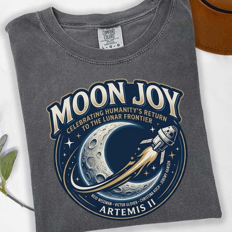 May include: A gray Comfort Colors t-shirt featuring a circular graphic with the words "MOON JOY" and "ARTEMIS II". The graphic depicts a crescent moon, a rocket, and the text "CELEBRATING HUMANITY'S RETURN TO THE LUNAR FRONTIER".