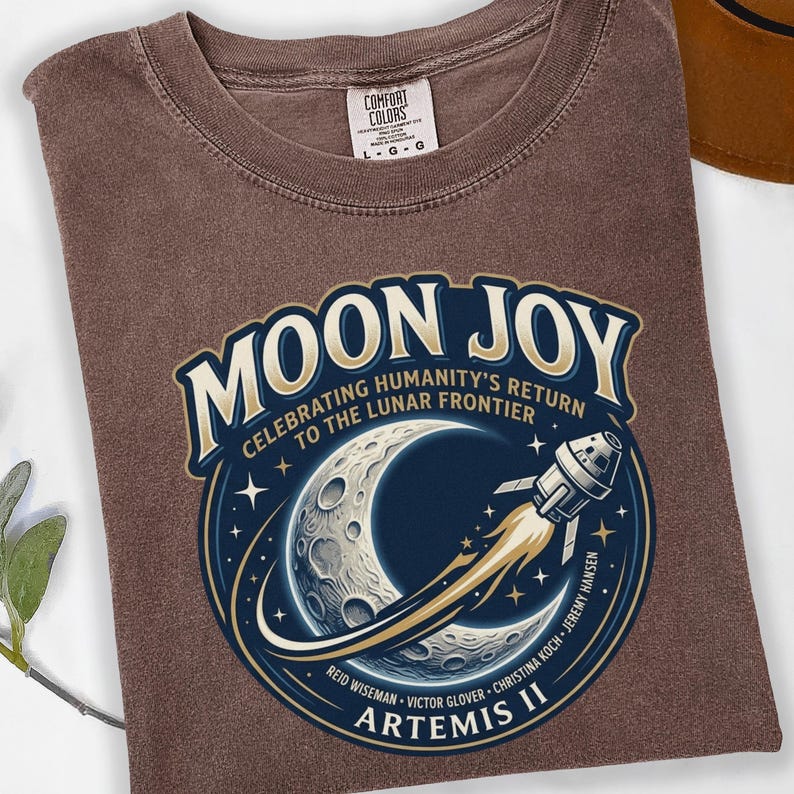 May include: A brown t-shirt featuring a circular graphic with the words "MOON JOY" and "CELEBRATING HUMANITY'S RETURN TO THE LUNAR FRONTIER". The graphic includes a crescent moon, a rocket, and the text "ARTEMIS II".