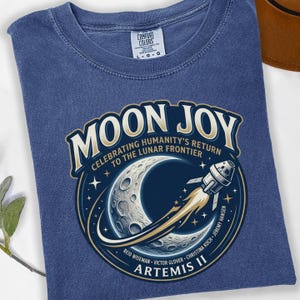 May include: A blue Comfort Colors t-shirt featuring a circular graphic with the words "MOON JOY" and "CELEBRATING HUMANITY'S RETURN TO THE LUNAR FRONTIER." The graphic includes a crescent moon, a rocket, and the text "ARTEMIS II."