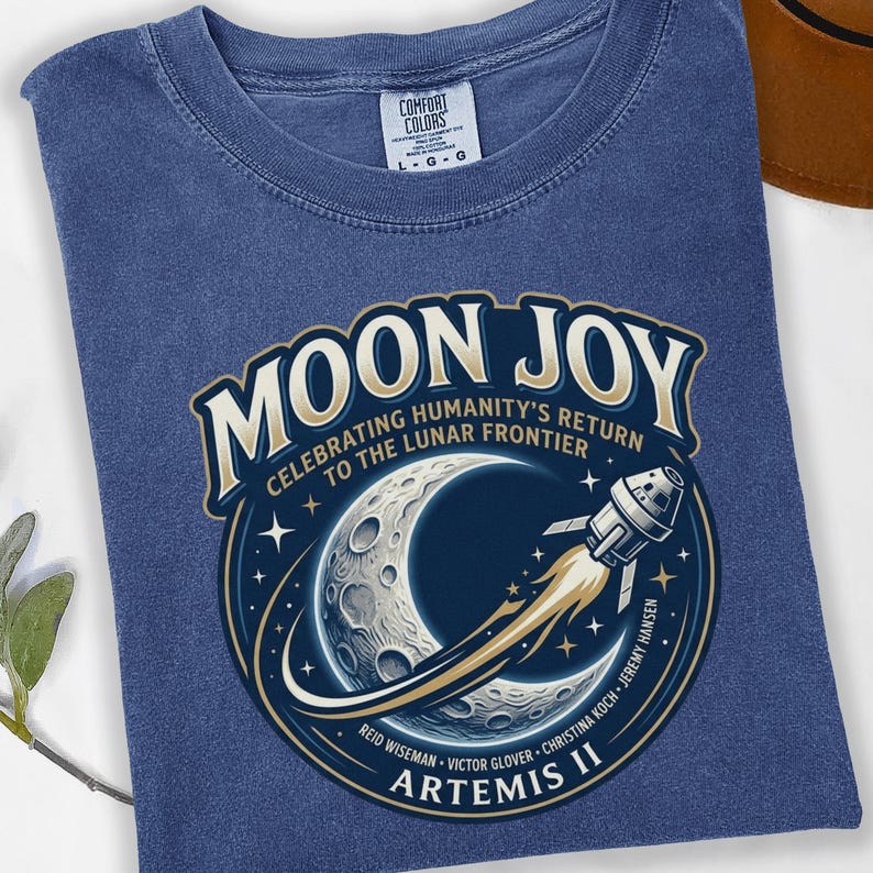 May include: A blue Comfort Colors t-shirt featuring a circular graphic with the words "MOON JOY" and "CELEBRATING HUMANITY'S RETURN TO THE LUNAR FRONTIER." The graphic includes a crescent moon, a rocket, and the text "ARTEMIS II."