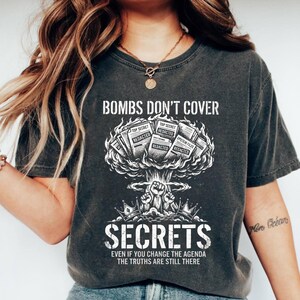 May include: Dark gray t-shirt with the text "BOMBS DON'T COVER SECRETS" and a graphic of a mushroom cloud with files labeled "Top Secret" and "Redacted." The bottom of the shirt reads "EVEN IF YOU CHANGE THE AGENDA THE TRUTHS ARE STILL THERE."