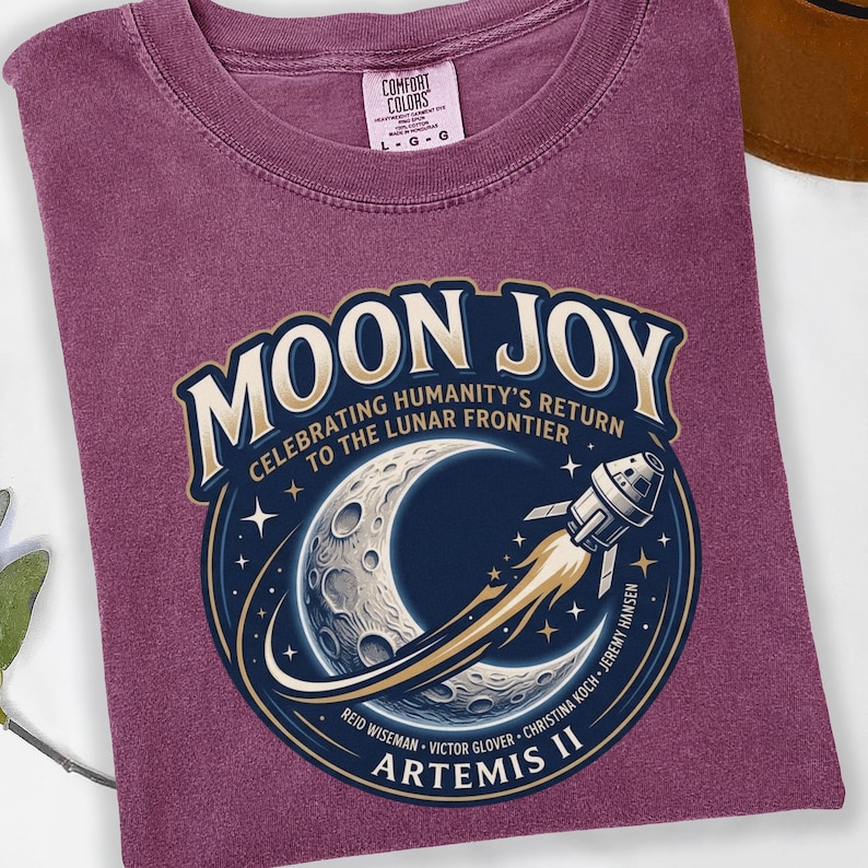 May include: A maroon Comfort Colors t-shirt featuring a circular graphic with the words "MOON JOY" and "CELEBRATING HUMANITY'S RETURN TO THE LUNAR FRONTIER." The graphic includes a crescent moon, a rocket, and the text "ARTEMIS II."