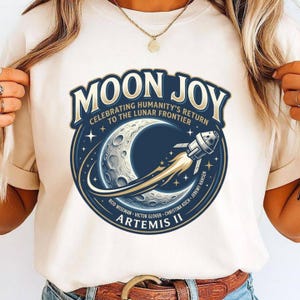 May include: Cream-colored t-shirt featuring a circular graphic with the words "MOON JOY" and "CELEBRATING HUMANITY'S RETURN TO THE LUNAR FRONTIER." The design includes a crescent moon, a rocket, and the text "ARTEMIS II."
