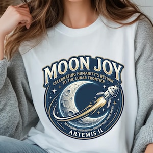 May include: White crew neck t-shirt featuring a circular graphic with the text "MOON JOY" and "CELEBRATING HUMANITY'S RETURN TO THE LUNAR FRONTIER". The design includes a crescent moon, a rocket, and the text "ARTEMIS II".
